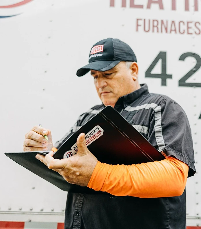 Man wearing a cap and uniform writes on a clipboard in front of an Alexander's Heating truck.